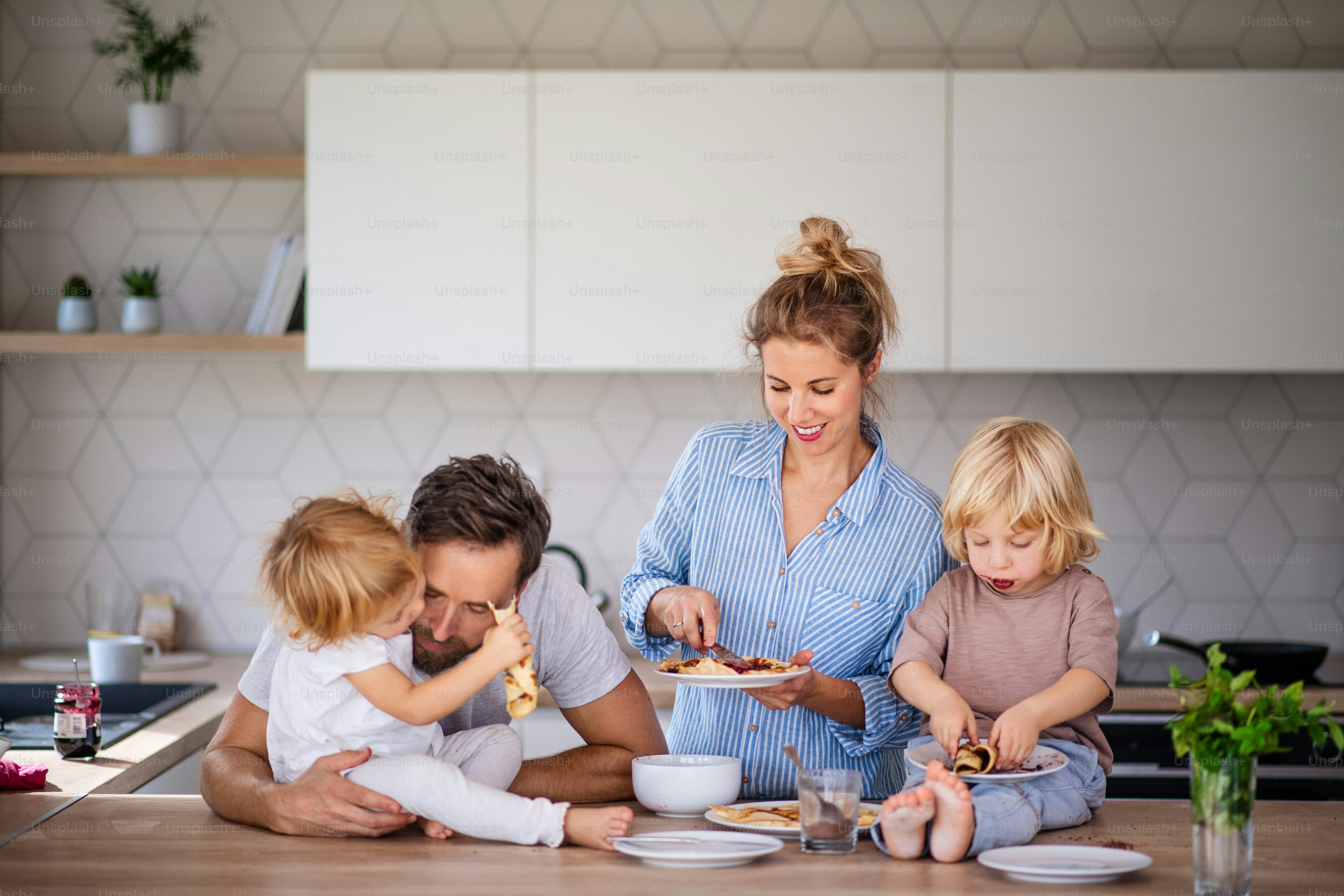 Front view of young family with two small children indoors in kitchen, eating pancakes